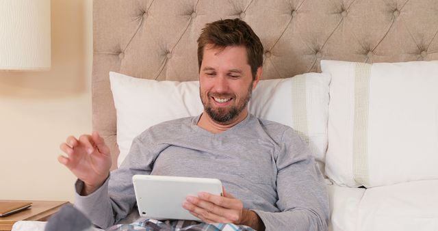 Middle-aged man in casual clothing smiling while using tablet in bed. Perfect for ads and articles about technology use, relaxation, home comfort, or leisure activities.