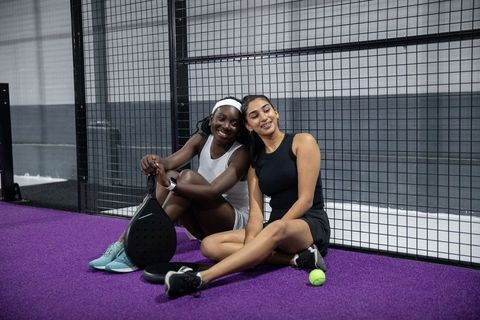 Smiling diverse women relaxing on purple padel court holding paddles and neon ball