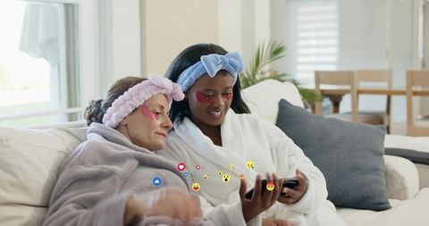 Relaxing friends in plush robes sharing phone and facial patches on cozy living room sofa