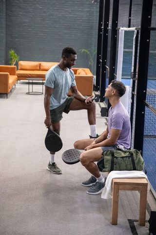 Diverse male friends resting after padel match, chatting courtside with gear and duffel