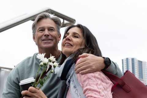 Middle-aged couple hugging on city balcony holding white flowers and takeaway coffee