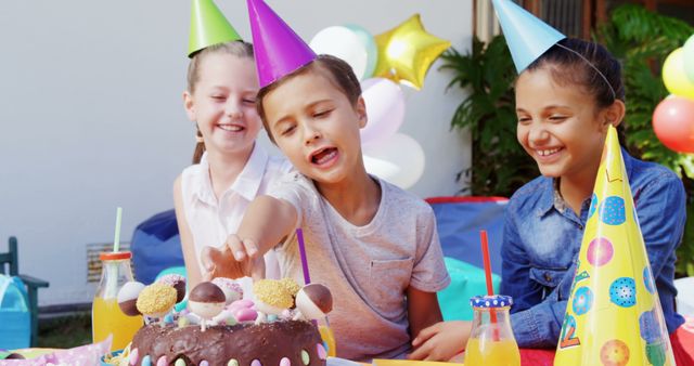A group of diverse children is celebrating a birthday party outdoors, with one child reaching for the cake, with copy space. Their joyful expressions and party hats add to the festive atmosphere of the occasion.