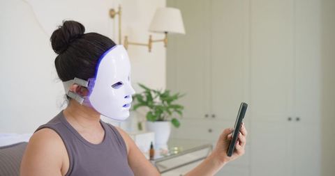 Woman wearing led facial mask using smartphone for home skincare and beauty routine