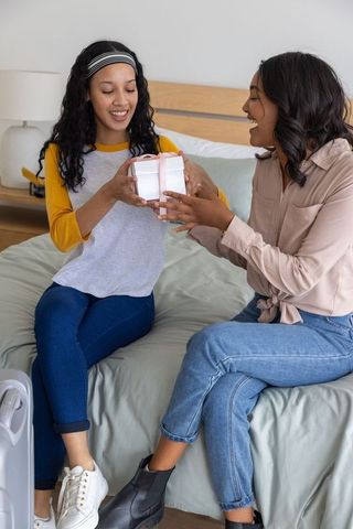 Diverse mother and teenage daughter exchanging gift, sharing joyful bonding moment on bed