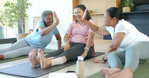 African american senior women laughing, high-fiving and stretching together in bright home yoga clas