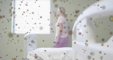 Mature woman adjusting blinds in hospital room with airborne dust particles and calm light