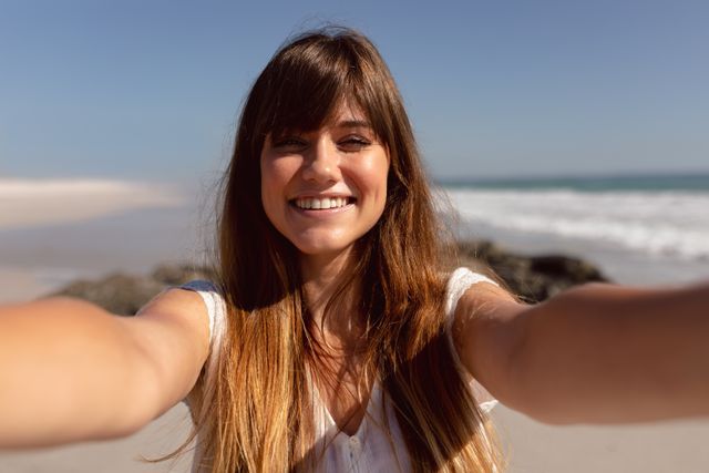 Young woman smiling and taking a selfie on a sunny beach. Perfect for use in travel blogs, vacation advertisements, lifestyle magazines, and social media promotions highlighting summer fun and outdoor activities.