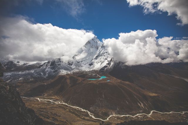 Image showcases majestic snow-covered mountain range partially hidden by clouds, with a river flowing below under a dramatic blue sky. Perfect for travel, adventure, nature-related marketing materials, and as a scenic backdrop in brochures or websites showcasing wilderness and outdoor activities.
