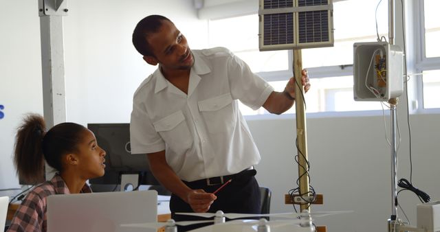An engineer in professional attire talks to a female student, demonstrating a solar energy system mounted on a pole. Both are in a well-lit room with technological equipment. Ideal for use in educational materials, articles about renewable energy, engineering programs, and classroom learning environments.