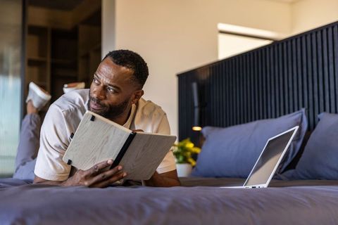 African american man reading notebook on bed with laptop in cozy modern home workspace