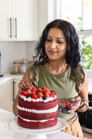 Woman decorating red velvet layer cake with strawberries and blackberries in sunlit kitchen