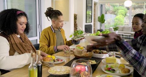 Diverse friends passing serving bowl and sharing cozy home dinner around wooden dining table