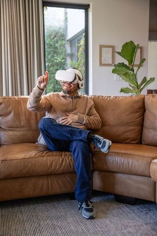 Middle-aged man using VR headset on leather sofa in modern living room with natural light
