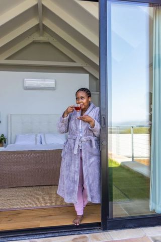 African american woman sipping tea in bathrobe on balcony with sunlit bedroom and ocean view