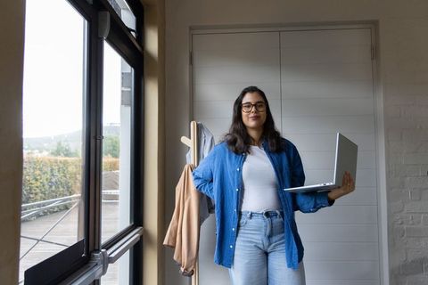 Asian woman standing in hallway holding laptop, working from home in casual denim