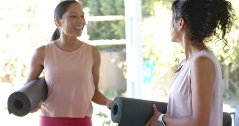 Two women carrying rolled yoga mats and chatting in sunlit studio by glass doors