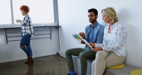 Colleagues reviewing color print while collaborating on bench in modern daylight office