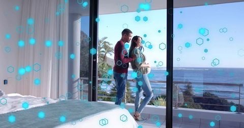 Expectant couple standing on seaside balcony overlooking ocean through bedroom glass door