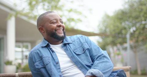 African american man smiling on patio bench in denim jacket, relaxing outdoors
