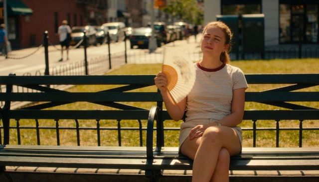 Young woman fanning herself on park bench in summer sunlight with urban street backdrop