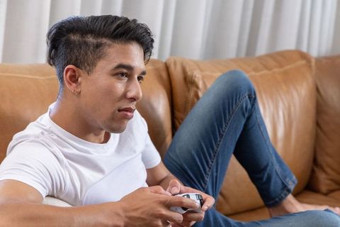African american man lounging on tan couch playing video game controller in white tee and jeans