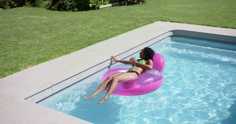 African american woman lounging on pink inflatable float in backyard pool holding drink