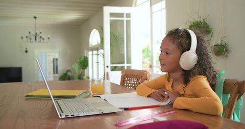 Young girl studying at home wearing headphones using laptop and notebook, bright natural light