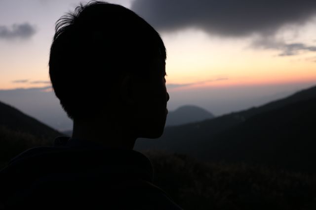 Boy's silhouette against a mountain landscape at sunset, evoking feelings of peace and contemplation. Ideal for articles about nature retreats, relaxation, mindfulness, solitude, travel destinations, and emotional introspection.