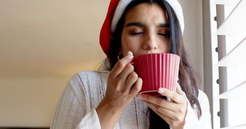 Festive woman in santa hat enjoying warm beverage by window