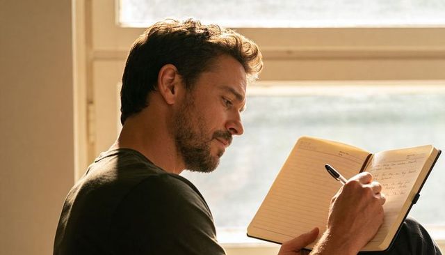 Bearded man writing in notebook by sunlit window, reflective journaling and quiet focus