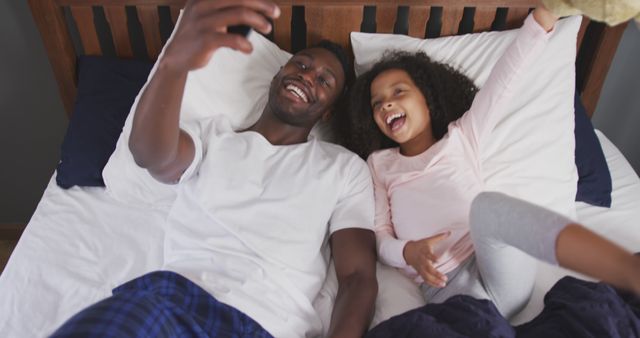 Happy african american father and daughter lying on bed taking selfies with smartphone. Fatherhood, childhood, fun, togetherness and domestic life.