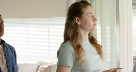 Diverse couple pausing at window during morning light, woman holding mug and gazing