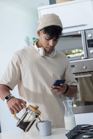 African-american man pouring french-press coffee while checking phone in modern kitchen