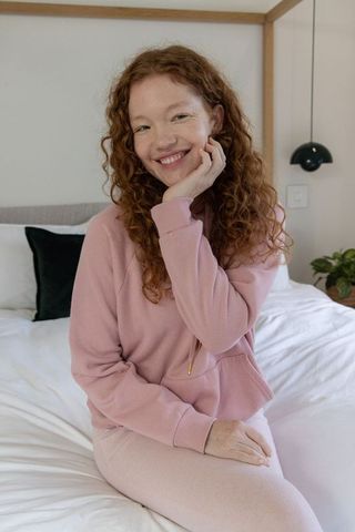 Smiling redhead woman sitting on bed wearing pink loungewear, cozy serene bedroom portrait