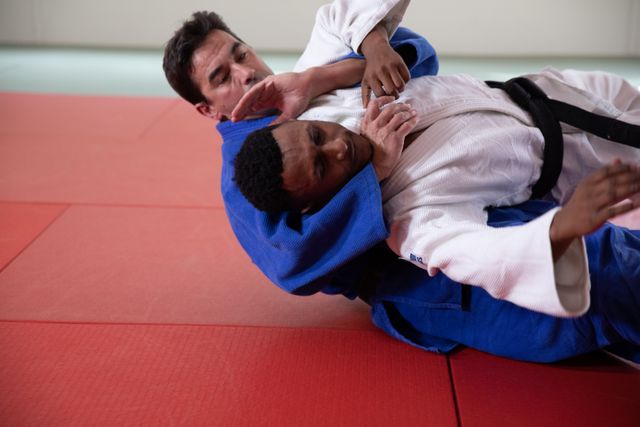 Caucasian and African American male judokas wearing blue and white judogi, practicing judo on a mat during a training in a bright studio.