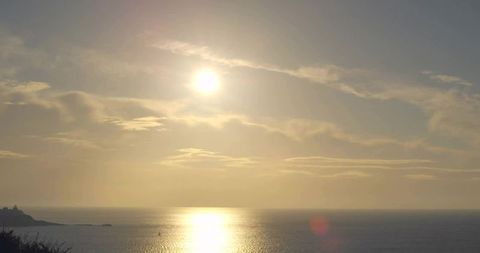 Golden sun reflecting on calm ocean horizon with layered clouds and rocky headland at golden hour
