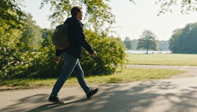 Man walking along sunlit park path carrying green backpack, casual urban nature stroll
