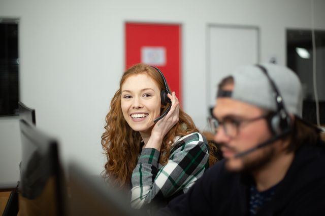 This image shows a happy woman with red hair working in a creative office environment, wearing a wireless headset and smiling at the camera. Ideal for use in articles or advertisements related to customer service, call centers, communication technology, teamwork, and professional office settings.