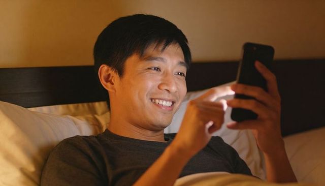 Smiling asian man relaxing in bed using smartphone at night, warm cozy intimate lifestyle