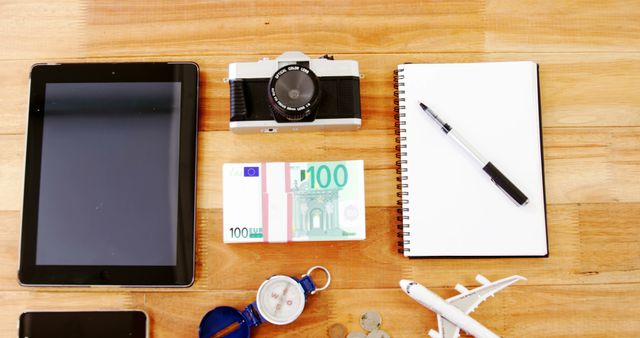 Flat lay arrangement of travel essentials on wooden table, including tablet, vintage camera, Euro currency notes, notebook, pen, compass, and small model airplane. Useful for travel blogs, packing tips articles, travel agency marketing, or vacation planning visuals.