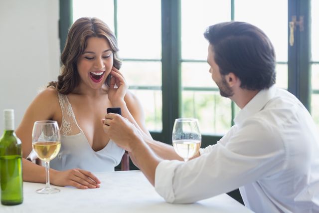 Man proposing to woman with engagement ring in restaurant. Woman looks surprised and happy. Wine glasses and bottle on table. Perfect for use in articles about proposals, romantic dining, relationships, and special moments.