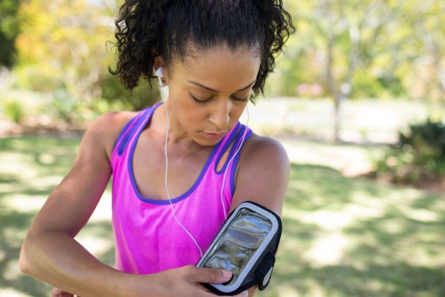 Woman in sportswear adjusting MP3 player in armband while jogging in park. Ideal for promoting fitness, outdoor activities, healthy lifestyle, and sports technology. Suitable for use in advertisements, fitness blogs, and health-related content.