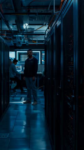 Vertical video: IT technician walking server aisle scanning blinking rack lights for maintenance