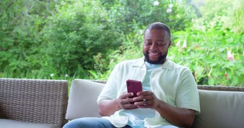African american man relaxing on patio sofa smiling while using smartphone in lush garden