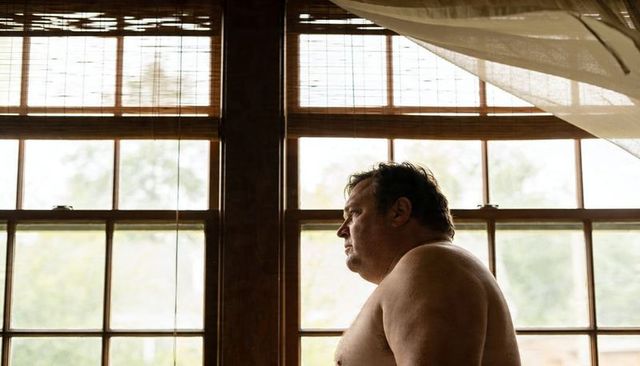 Contemplative shirtless man standing, gazing through soft backlit wooden windows with bamboo blinds