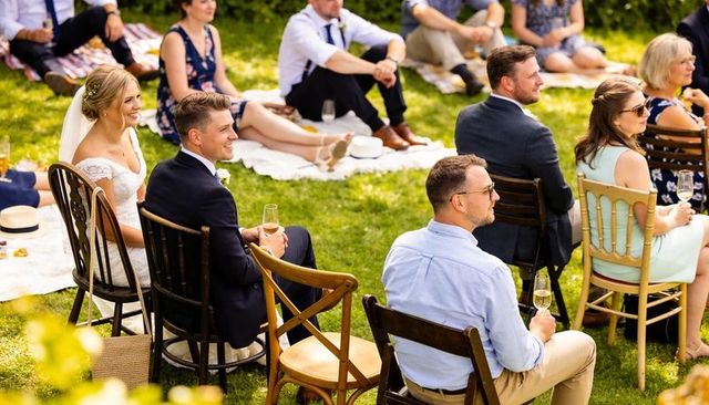 Sunlit outdoor wedding couple smiling and holding champagne flutes while seated on lawn