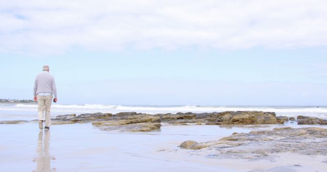 Senior man walking alone on rocky beach with clear sky and calm ocean. Ideal for themes of solitude, retirement, peaceful moments, nature walks, and healthy living.