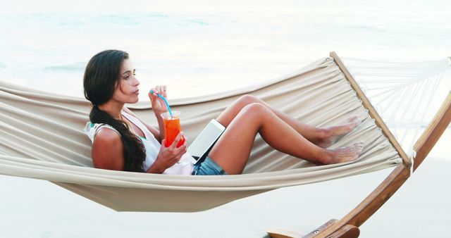 Scene of young woman relaxing in hammock by the tropical beach while sipping a cocktail and using a tablet. Ideal for content related to vacations, relaxed lifestyle, beach getaways, leisure activities, and summer holidays.