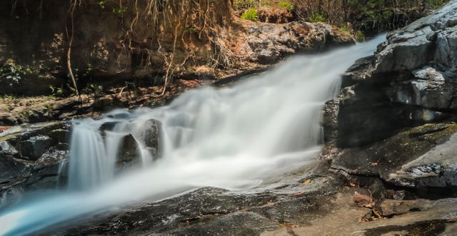 Image showcases a peaceful waterfall flowing over rocky terrain in a forest. Ideal for promoting nature conservation, outdoor activities, relaxation and travel destinations. Perfect for use in brochures, websites, and digital media related to natural beauty, environment, and tranquility.
