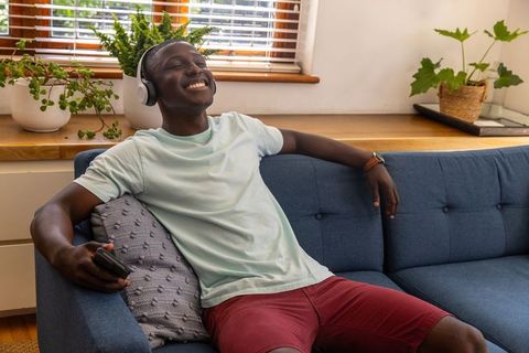 Relaxing African American man listening to music on couch with headphones and remote